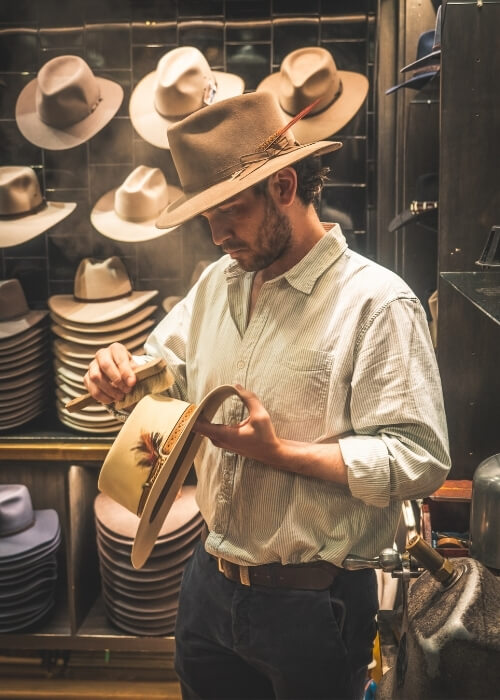 Hamish, the manager of Strand Hatters standing in the store brushing a hat.