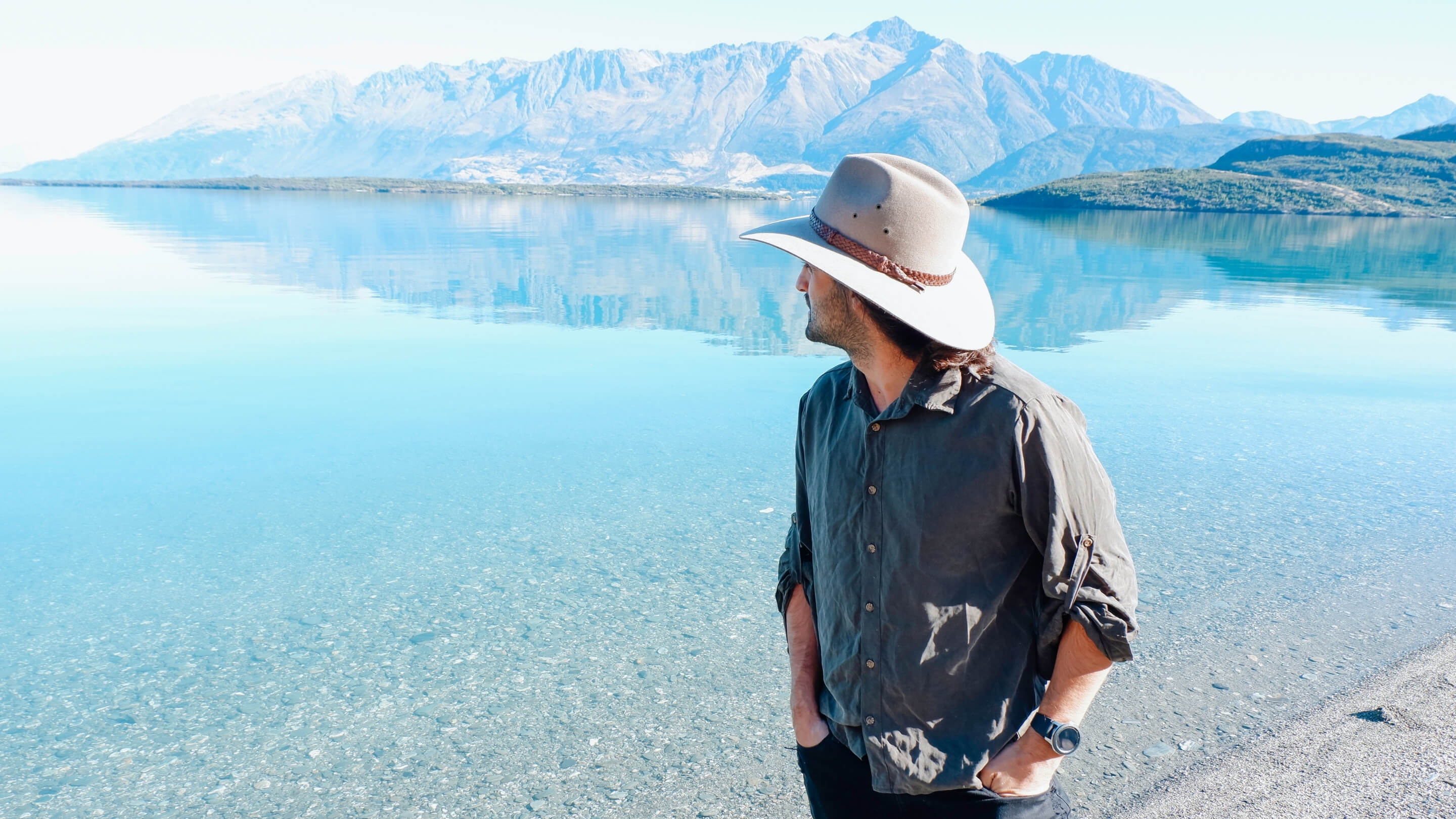Man wearing Akubra hat staring our across a lake with mountains in the background.