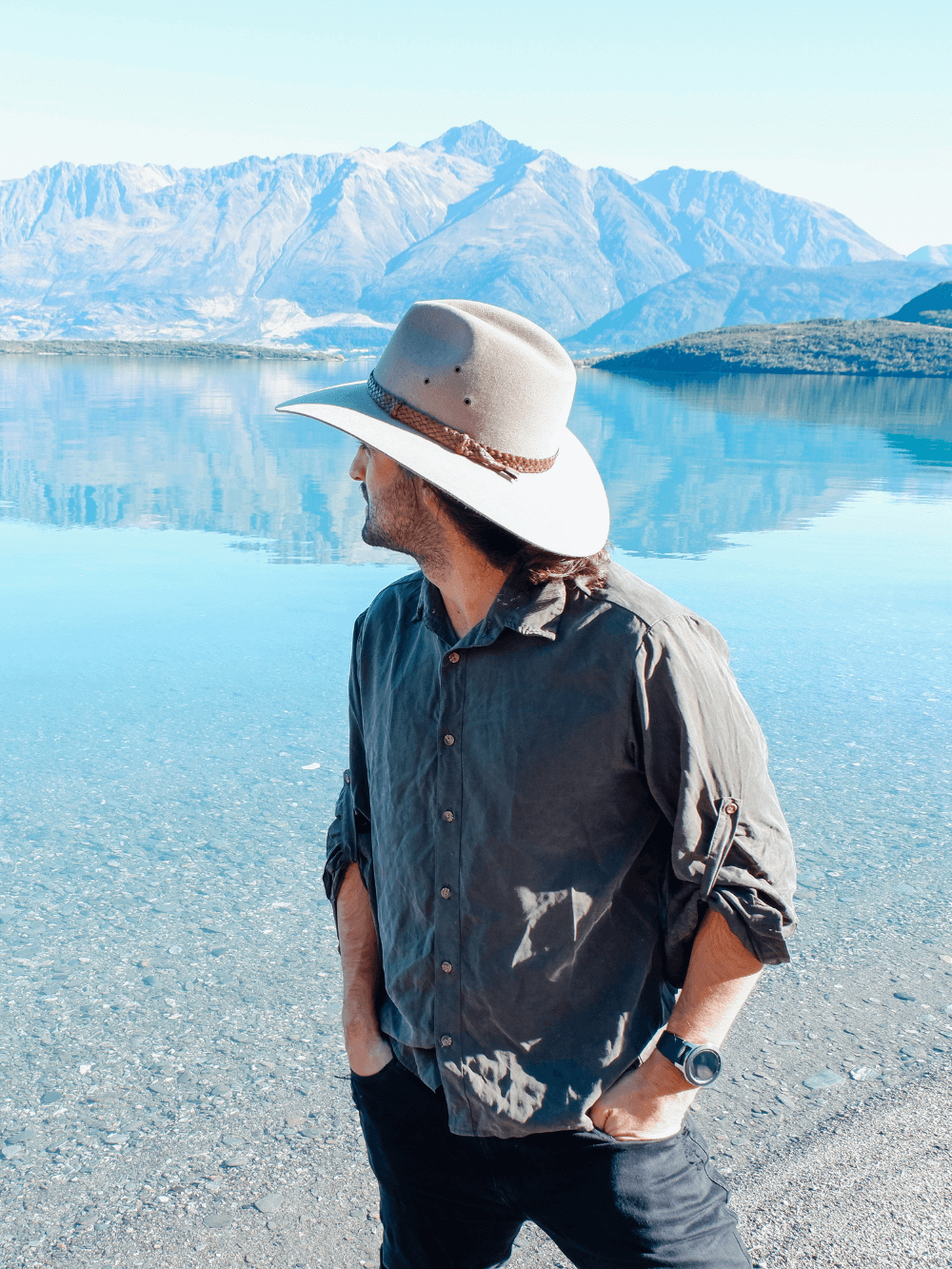 Man wearing Akubra hat staring our across a lake with mountains in the background.