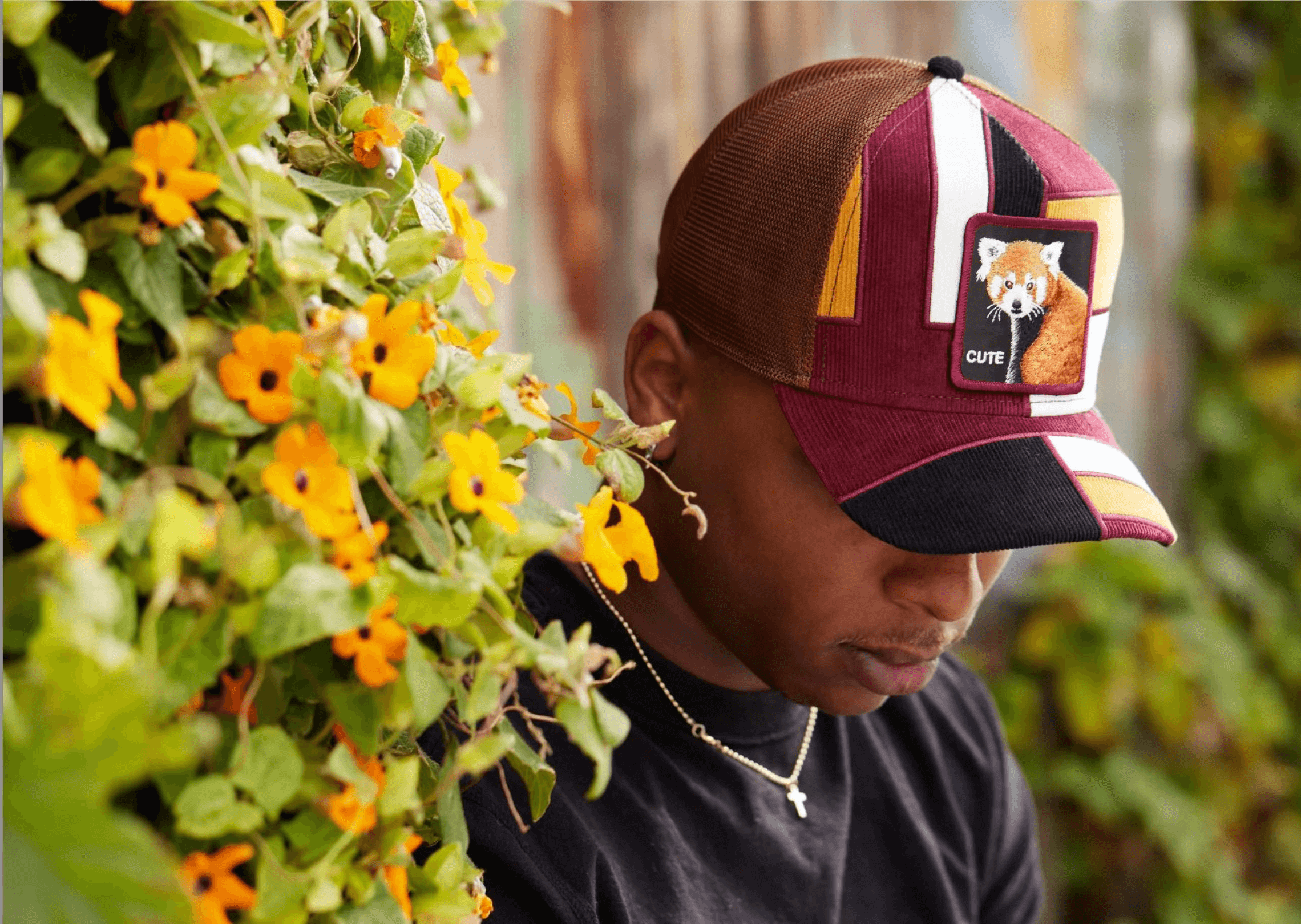 Man looking down, wearing Goorin Bros Trucker cap. He is standing next to a wall of flowers.